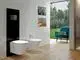 Weißes Wand-WC von Ideal Standard und eine Betätigungsplatte an einer hellen Wand. Im Hintergrund ein Fenster mit Blick auf einen gepflegten Garten und ein blauer Sessel.